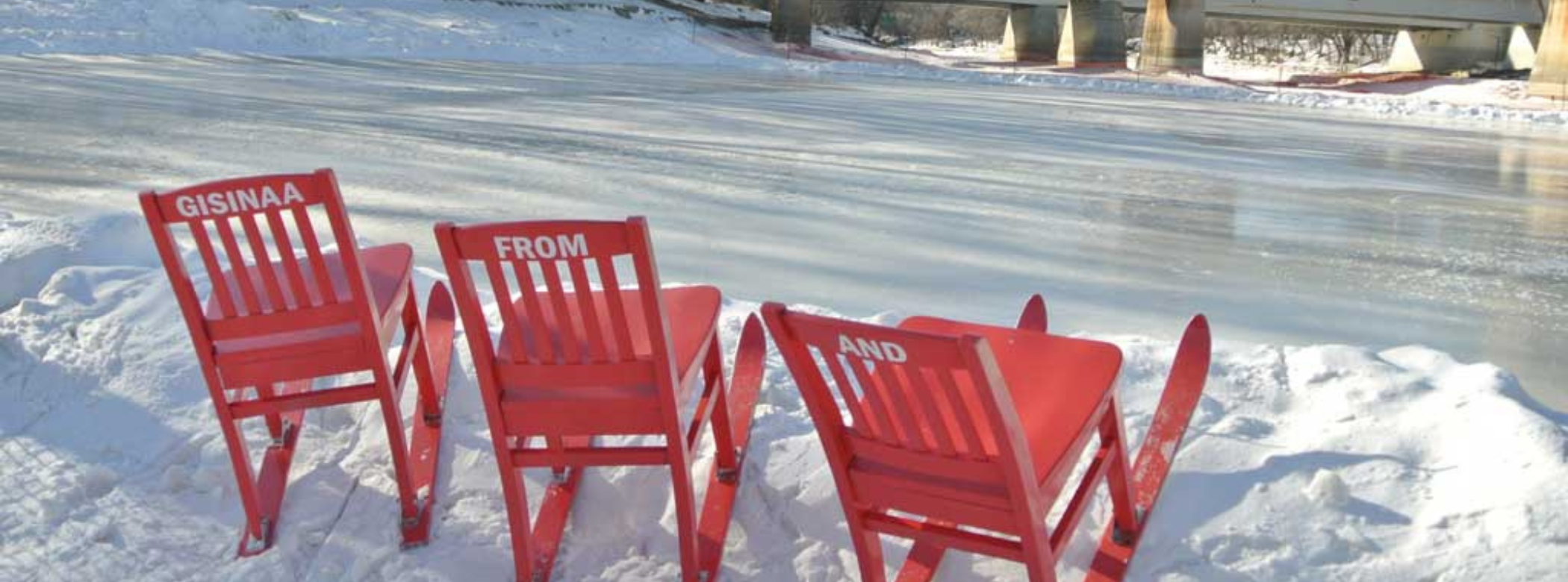 Three red chairs with skiis on their feet in a snowbank along the side of a cleared, frozen river. The backs of each chair has a word written on it in white, 