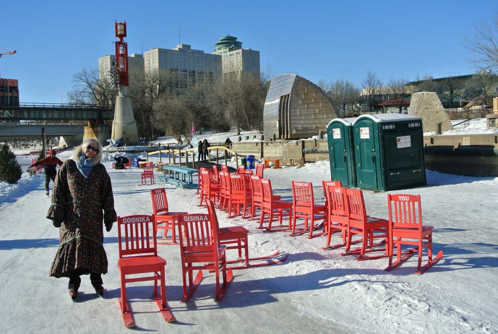 Maureen Matthews, bundled up in winter clothes, standing outside on a cleared forzen river path beside a number of red chairs with skiis along their feet. On the two chairs closest to her, the words "Goonika" and "Gisinaa" are written on the chair backs.