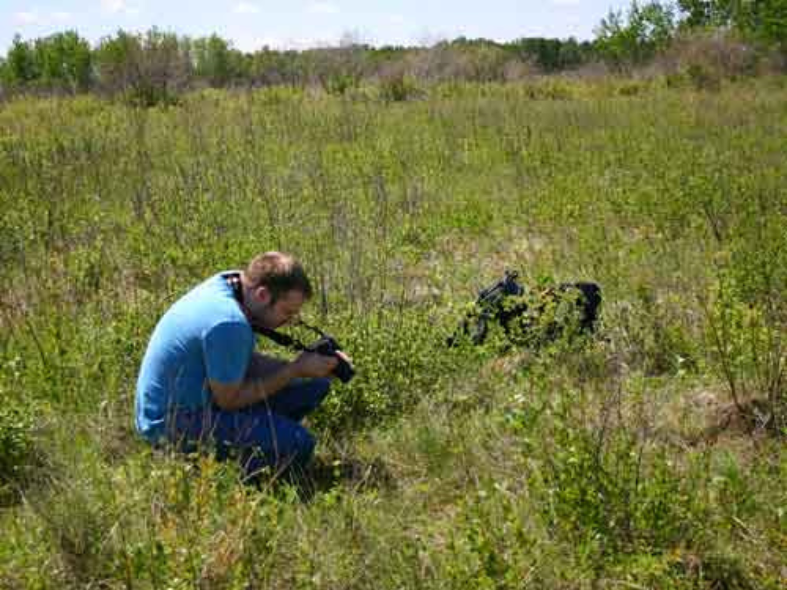 An individual crouched in a field with a DSLR camera, taking a photo of something in the field.