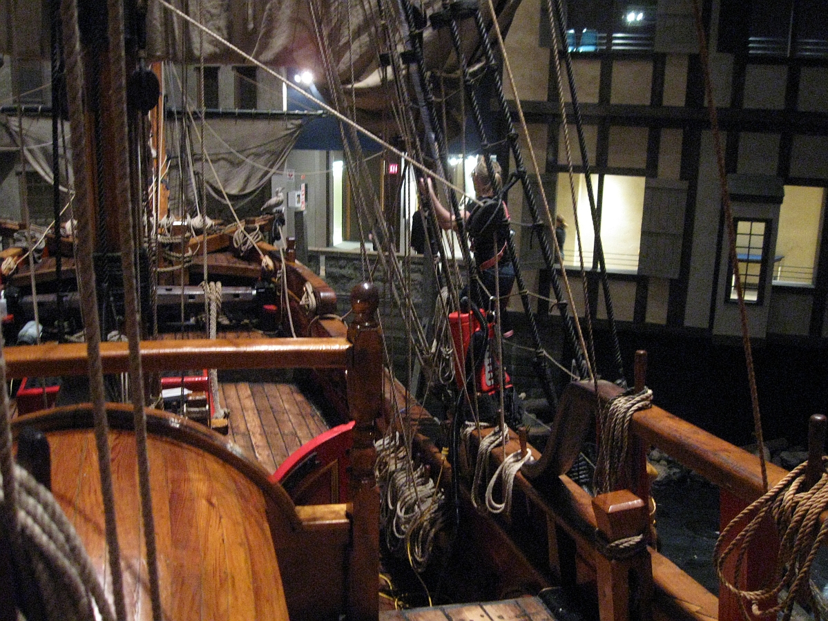 An individual in safety harness on one of the side rails of a wooden sailing vessel (the Nonsuch), preparing to climb up the ratlines with a backpack style vacuum cleaner.