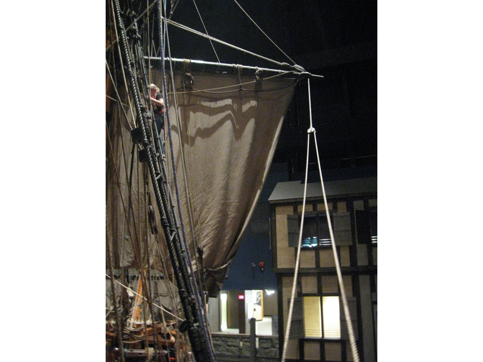 An individual in safety harness climbing the ratlines of a wooden sailing vessel (the Nonsuch) in full sail.