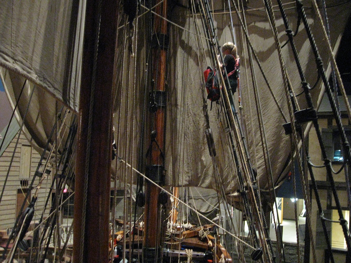 An individual in safety harness part way up the ratlines of a wooden sailing vessel (the Nonsuch) with a backpack style vacuum cleaner held in front of them through the ratlines.