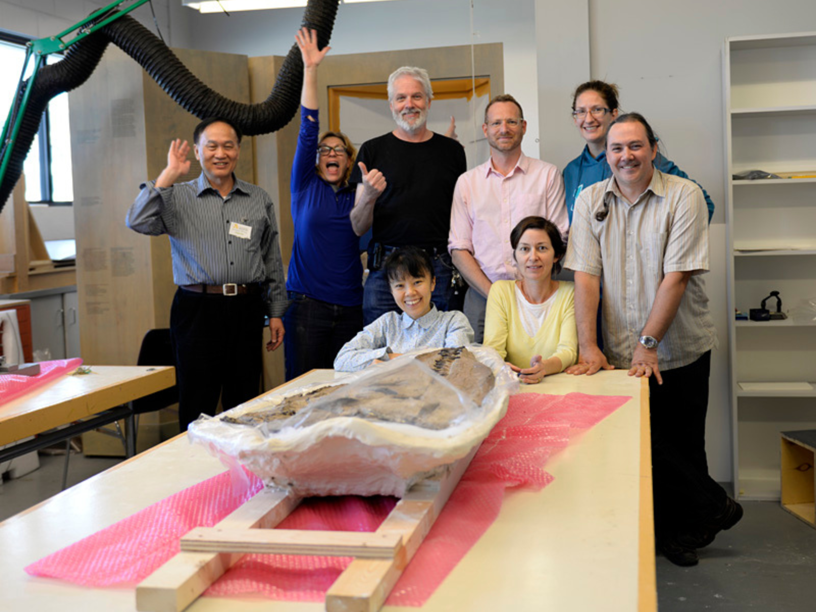 A group of eight people pose, smiling together, for a photo at the end of a table which has the flipped plesiosaur skull laid across it.