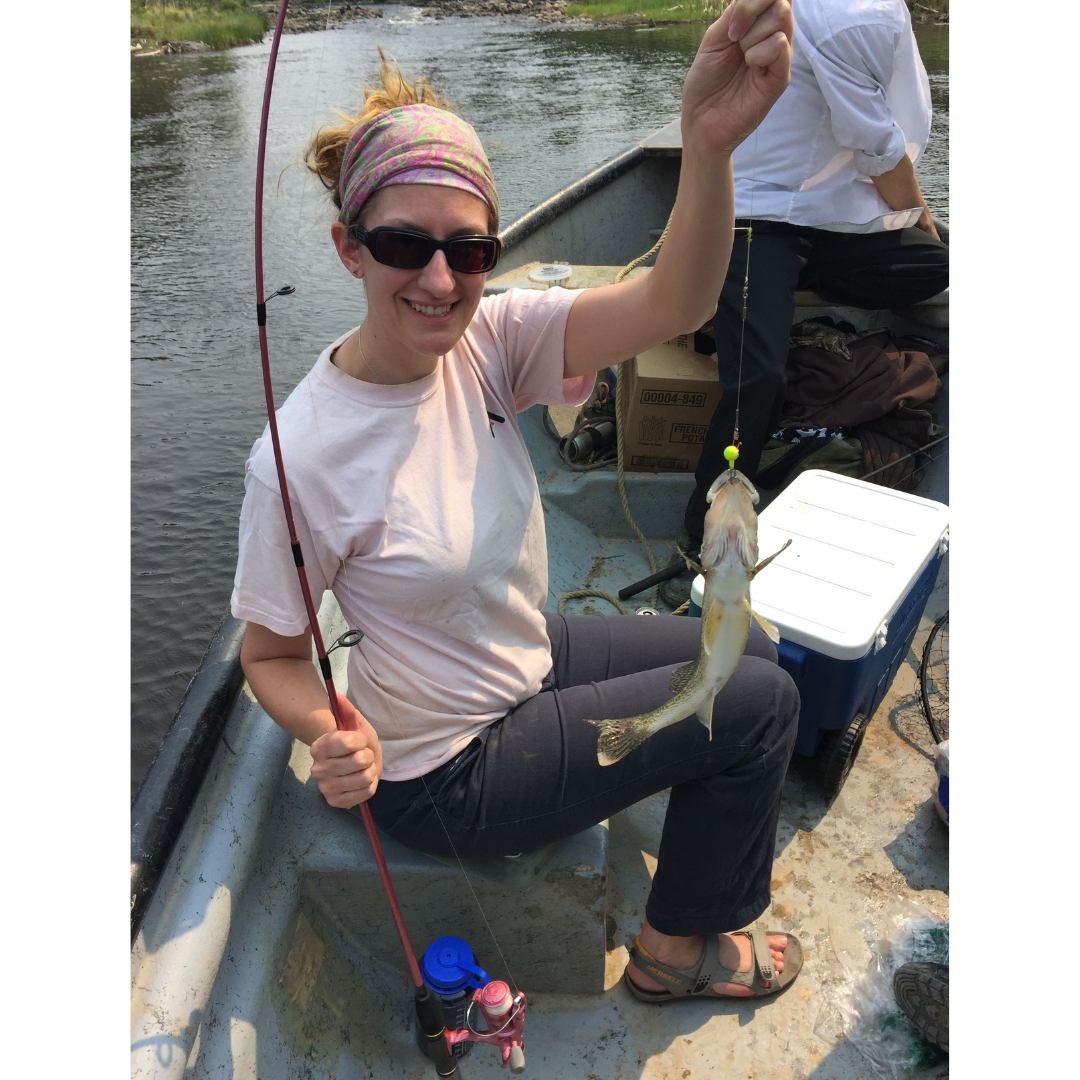 Dr. Amelia Fay seated in a small boat holding up a fish on the end of a fishing rod line.