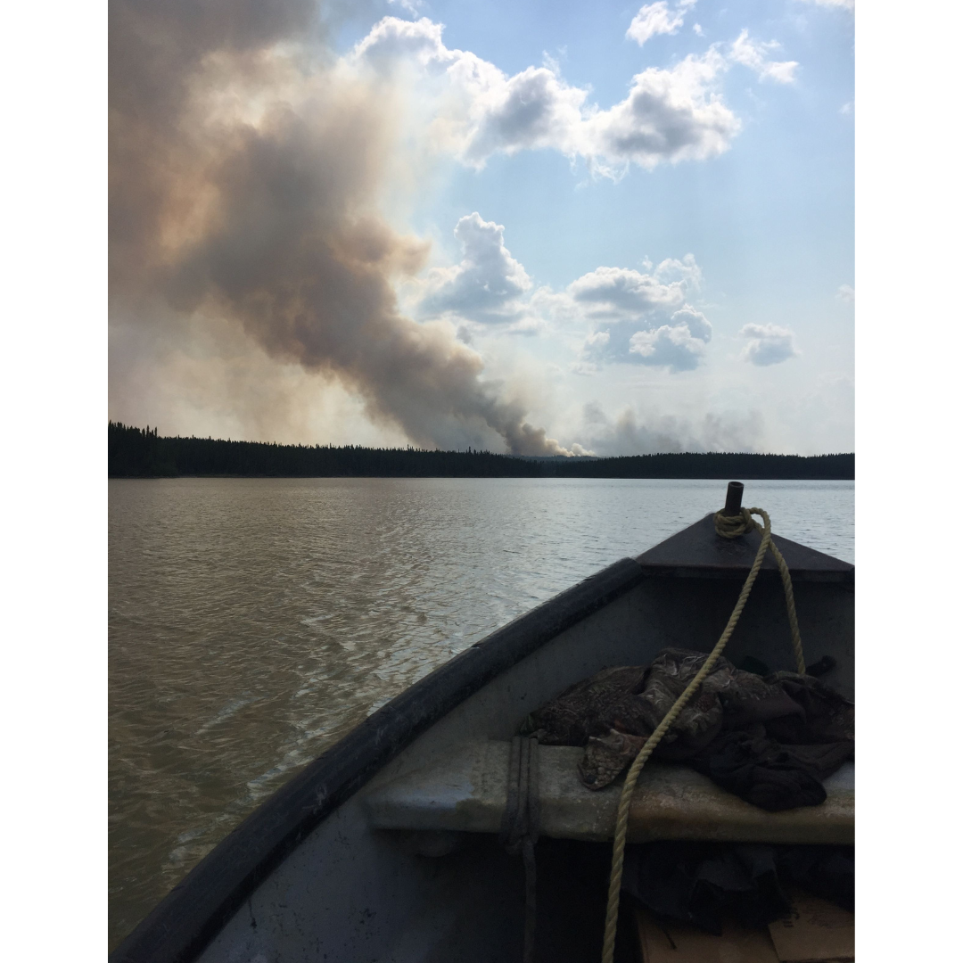 The front end of a small boat on a lake. A thick column of dark smoke rises from the distant shore.