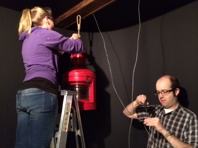 One individual on a ladder hanging a large ship's lantern for display, while another individual stands nearby working on some electrical wiring.