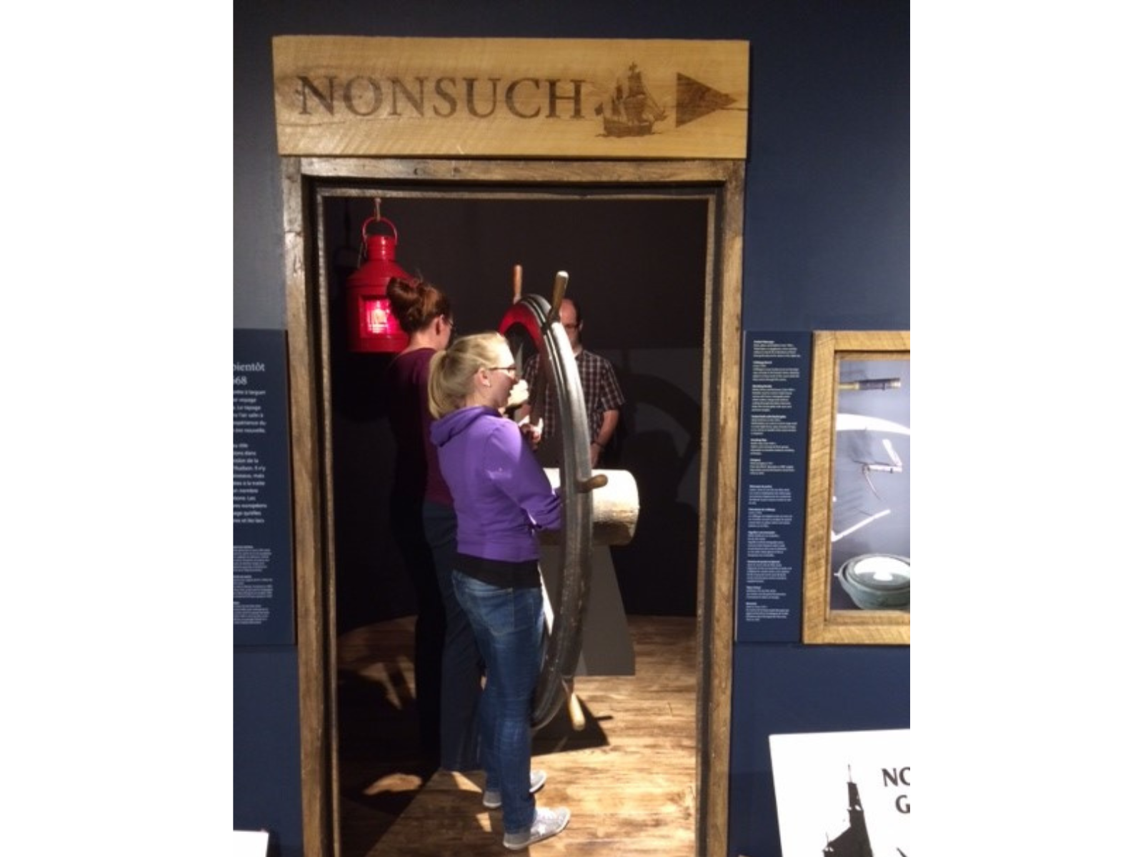 Three individuals moving together to transfer a large wooden ship's wheel into an exhibit case.