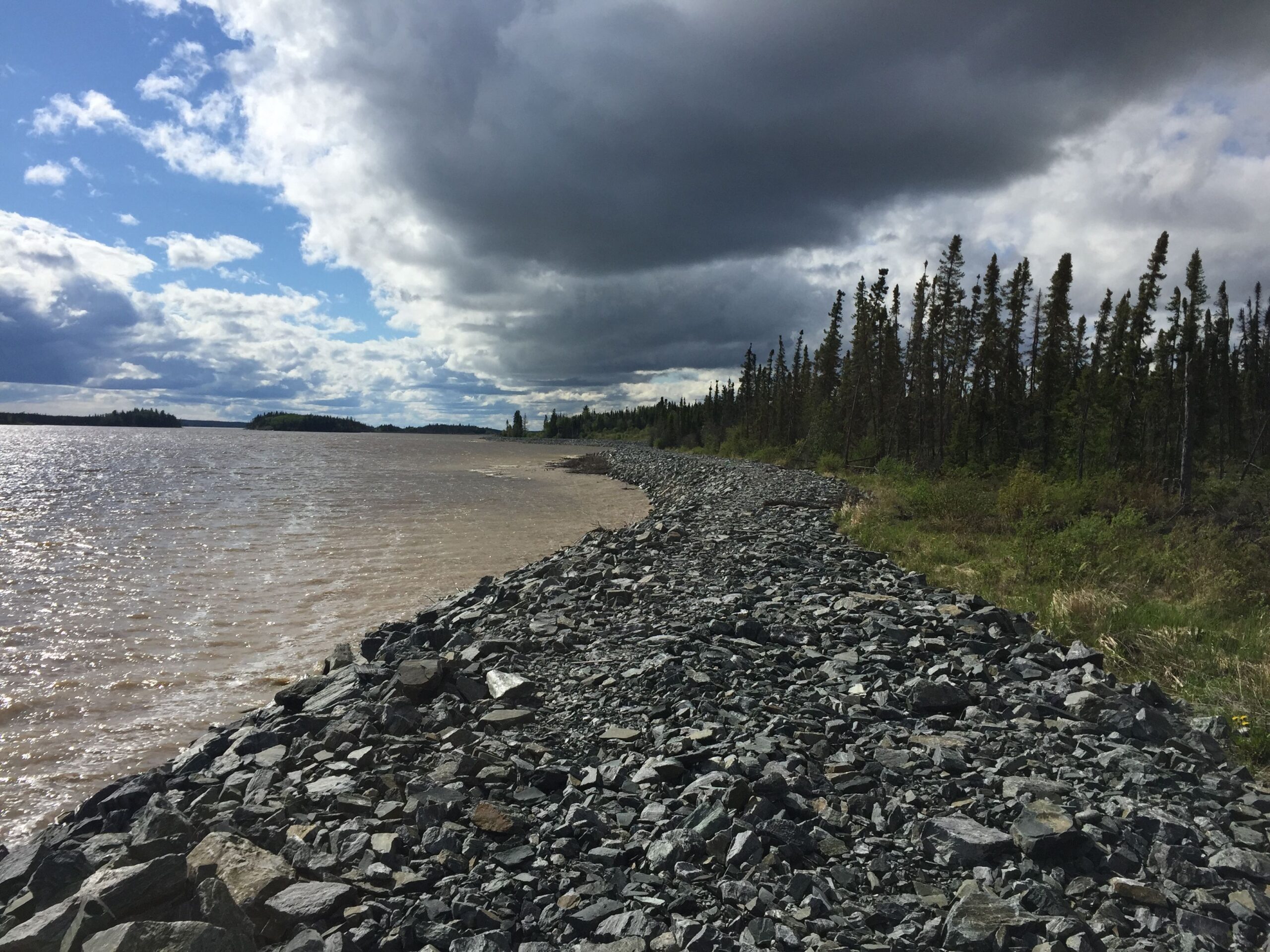 View down a rocky shoreline with dark clouds gathering overhead with the lake on one side and trees on the other.