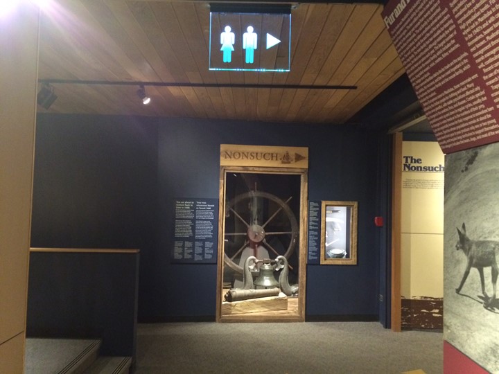 An open entryway with a two steps going up the the left, and a hallway to the right. Directly in front is an exhibit case with a large wooden steering wheel, ship's bell, and small canon on display. Signs overhead direct visitors right for both the Nonsuch and the bathrooms.