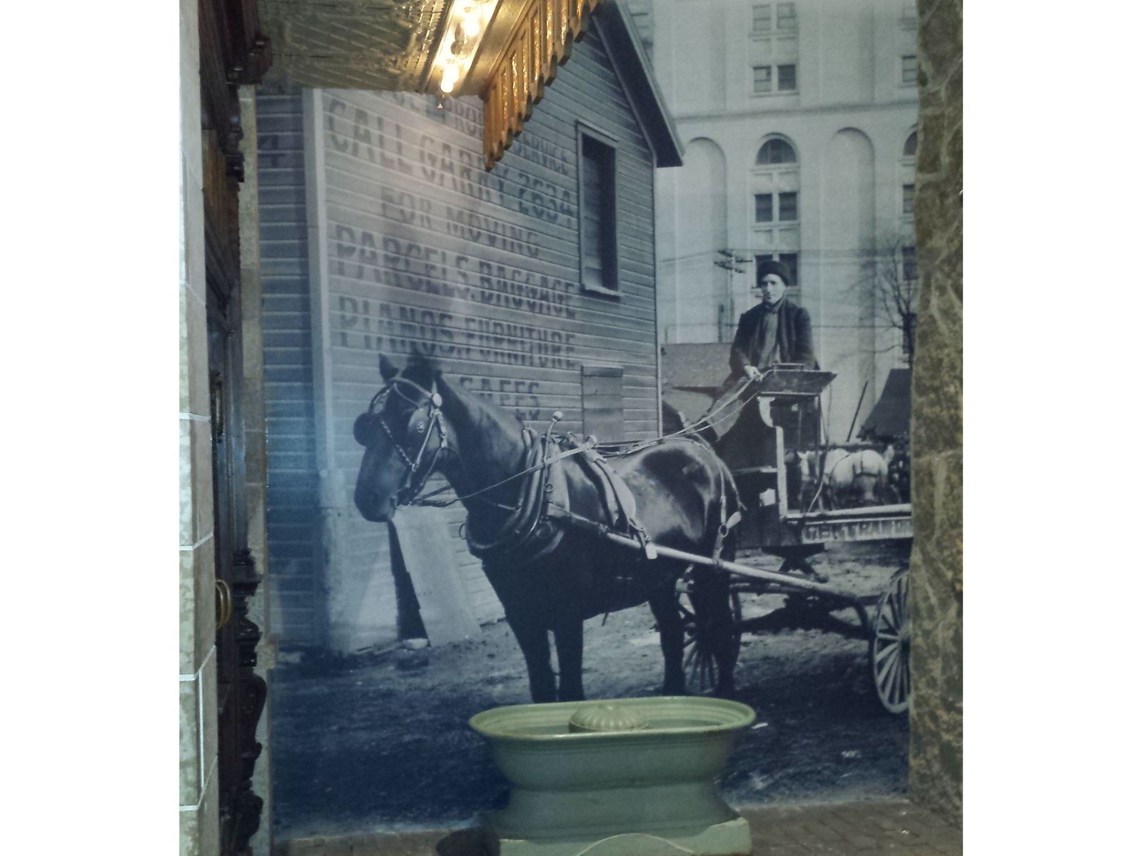 Historical photograph blown up between two stone walls. Photo shows a horse drawn wagon with a worker on the wagon with the reins. In front of the photo is a water trough.