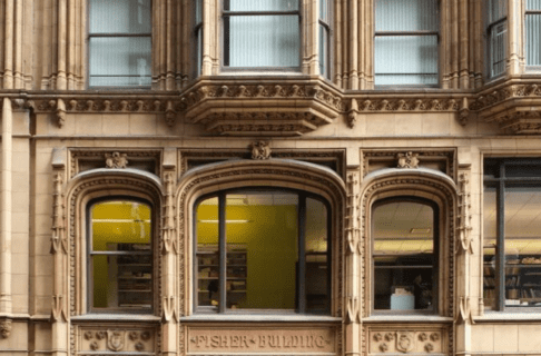 The front of a multistoried neo-Gothic building in sand-coloured stone with three large windows on each story.