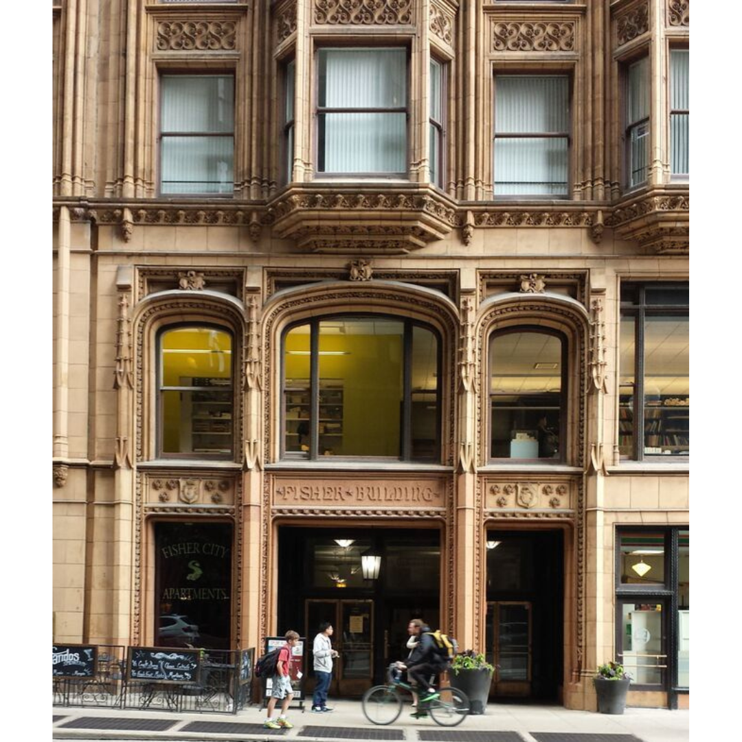 The front of a multistoried neo-Gothic building in sand-coloured stone with three large windows on each story. Pedestrians walk or bike past in front.