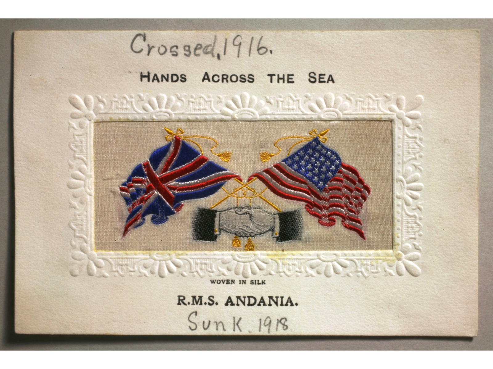 A postcard showing an embroidered image of two hands shaking under British and American flags. Printed text on the card frame reads, "Hands across the sea / R.M.S. Andania / Woven in silk". Handwritten text on the frame reads "Crossed, 1916 / Sunk 1918".