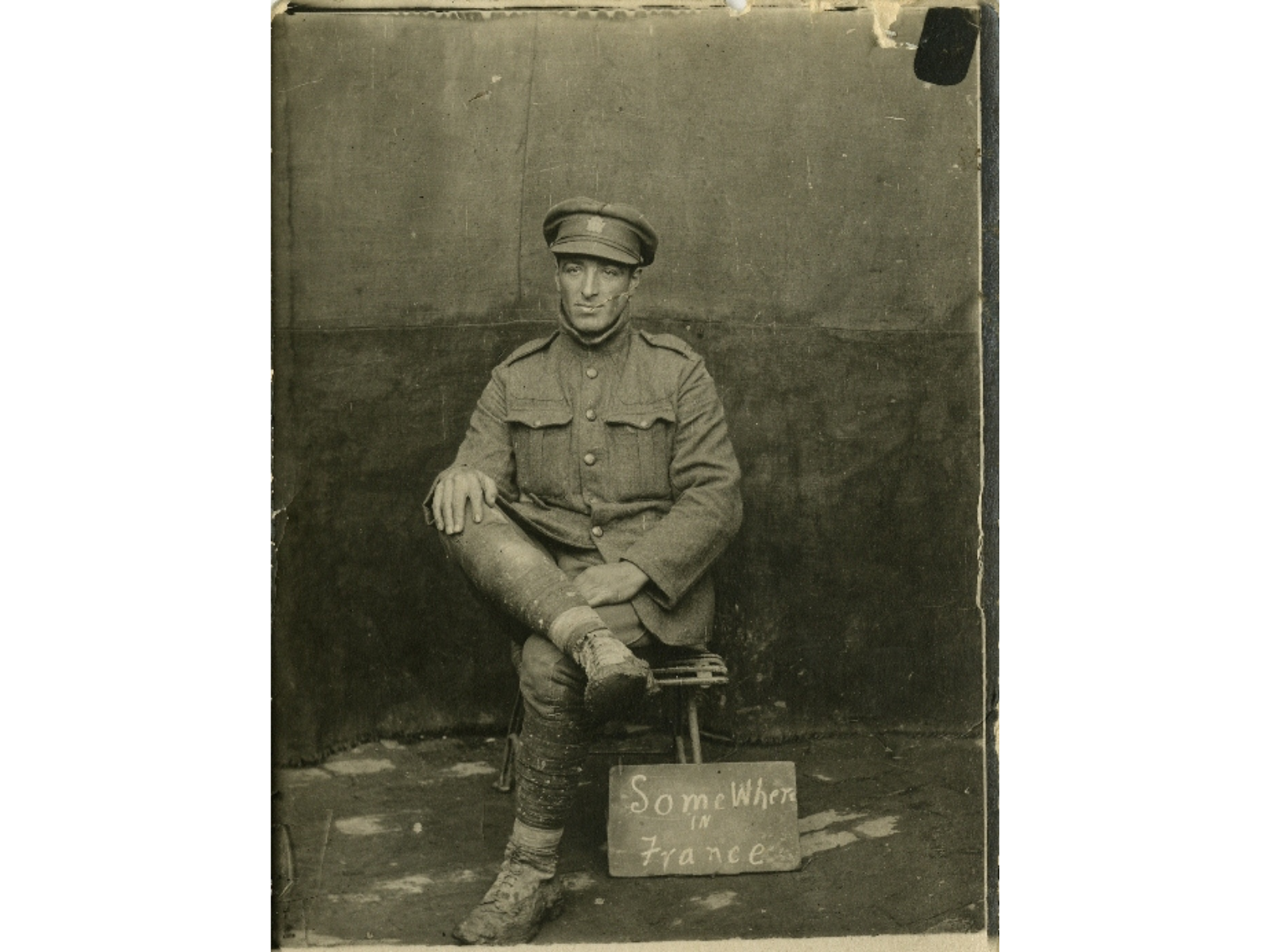 A sepia-toned photograph of a solider sitting on a stool with one leg up across the other. At the base of the stool and hand written sign reads, "Somewhere in France".