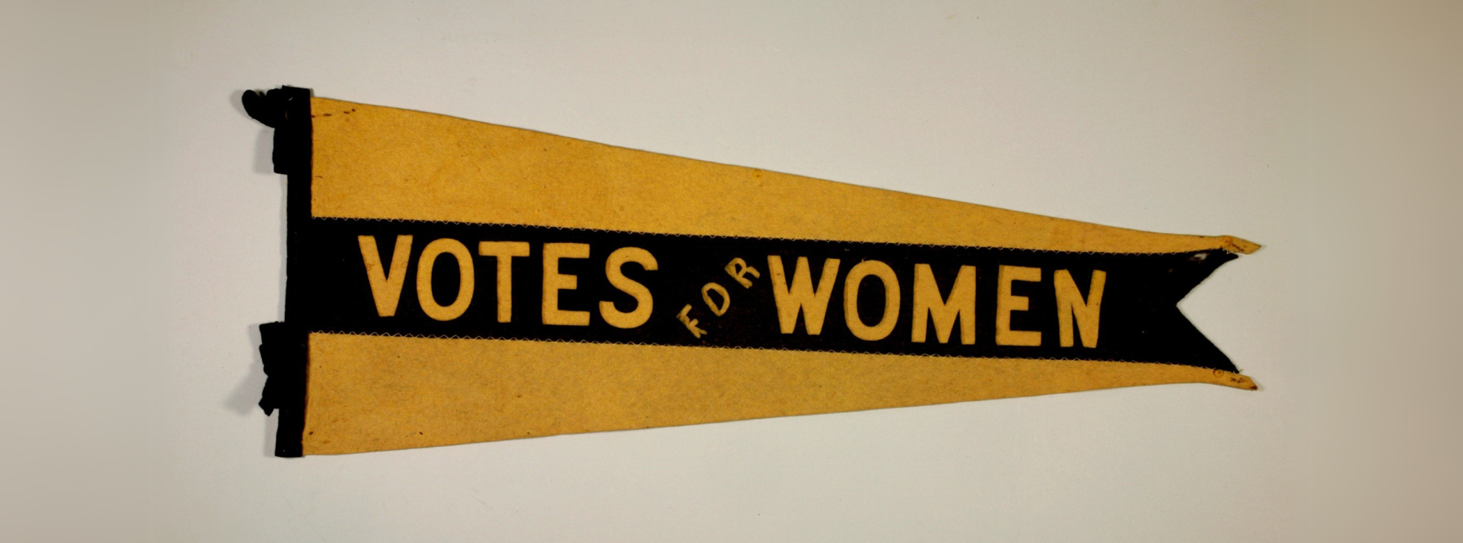 Elongated triangular flag. The background is yellow wiith a thick black stripe stitched on top reading “Votes for Women” along the centre.