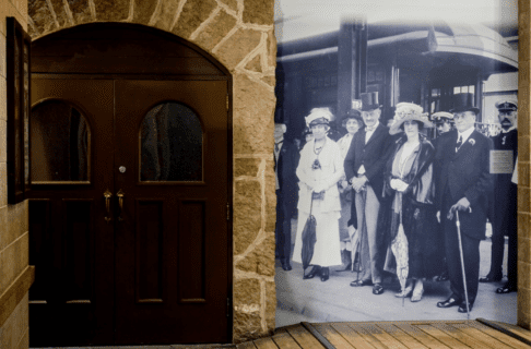 A historical photograph blown up on a stone wall next to a set of wooden doors. The photo shows a group of well dressed individuals from the 1920s standing at a rail station.