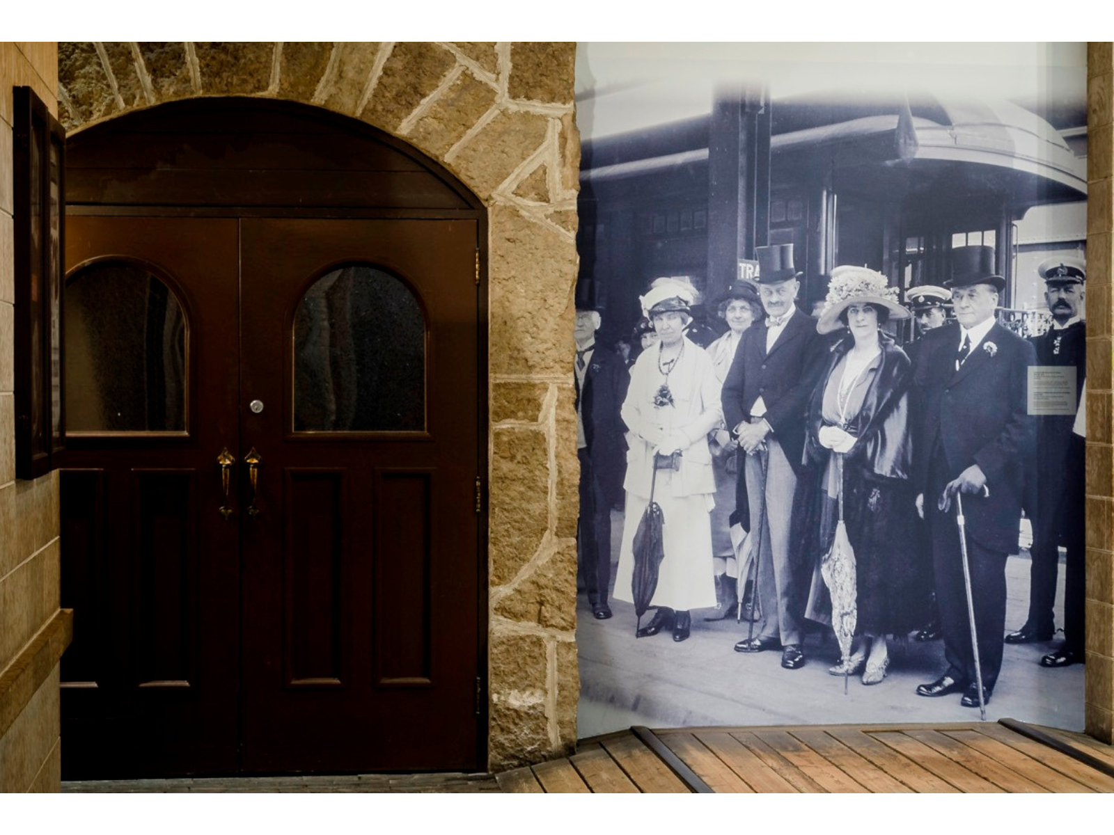 A historical photograph blown up on a stone wall next to a set of wooden doors. The photo shows a group of well dressed individuals from the 1920s standing at a rail station.