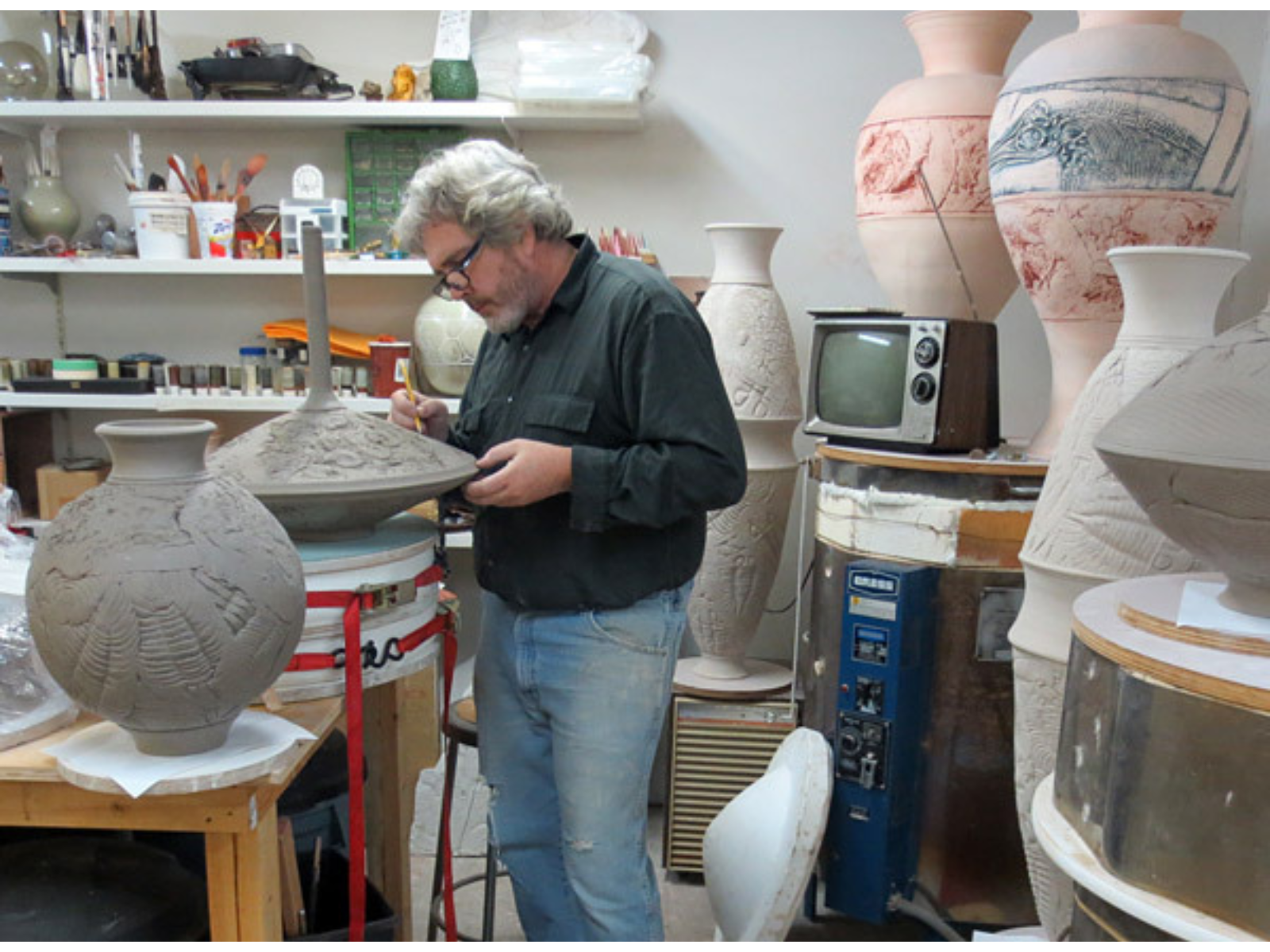 An individual engraving a ceramic in progress in a workshop with other partially completed ceramic vases.