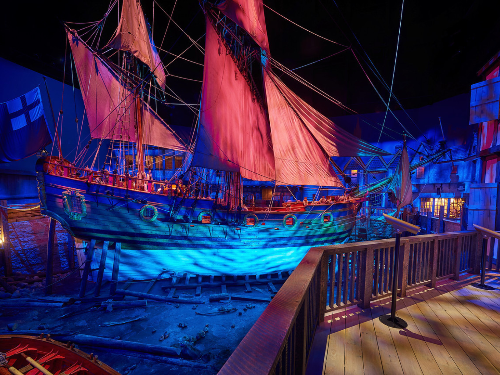 A large wooden sailing vessel in full sail in a museum gallery, dramatically lit with blue and red-orange lighting.