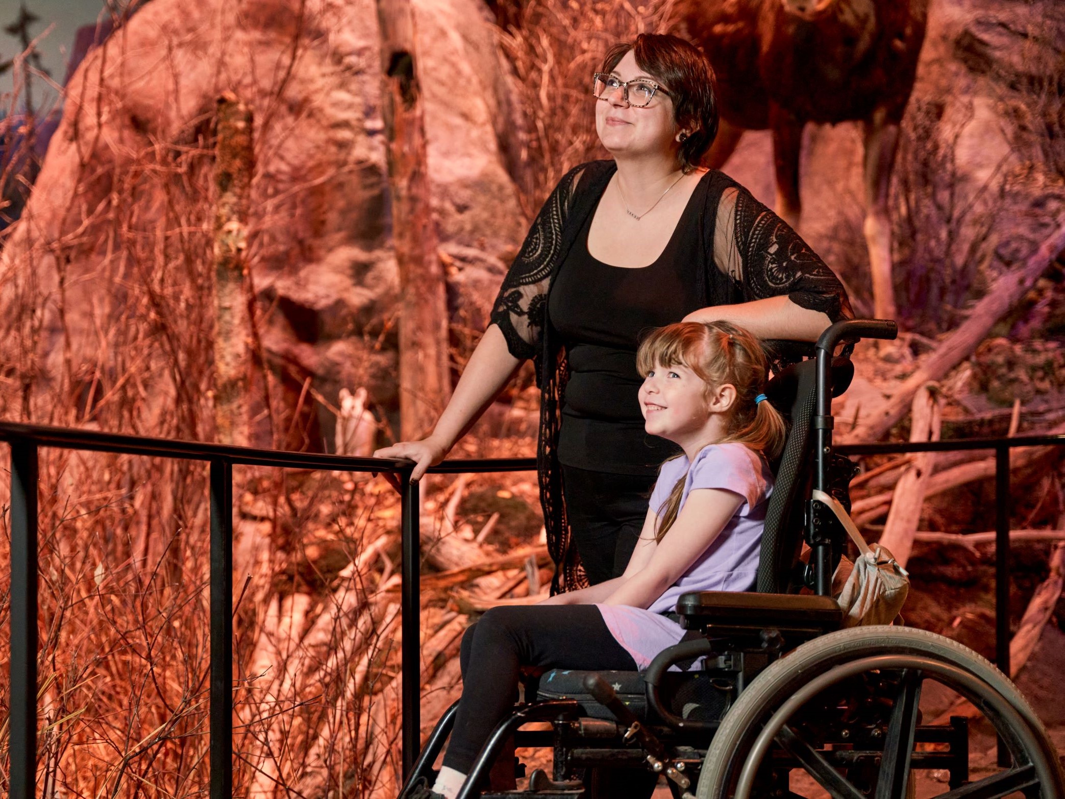 An adult standing beside a child in a wheelchair. Both smile as they look into the Manitoba Museum's immersive Boreal Forest diorama.