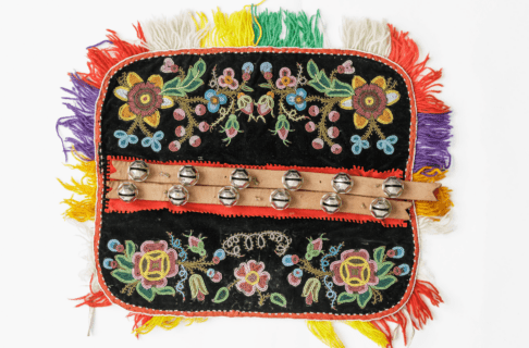 Close up on a beaded dog blanket. Detailed floral beading on a black background, with a colourful fringe along the edges, and two rows of bells down the centre.