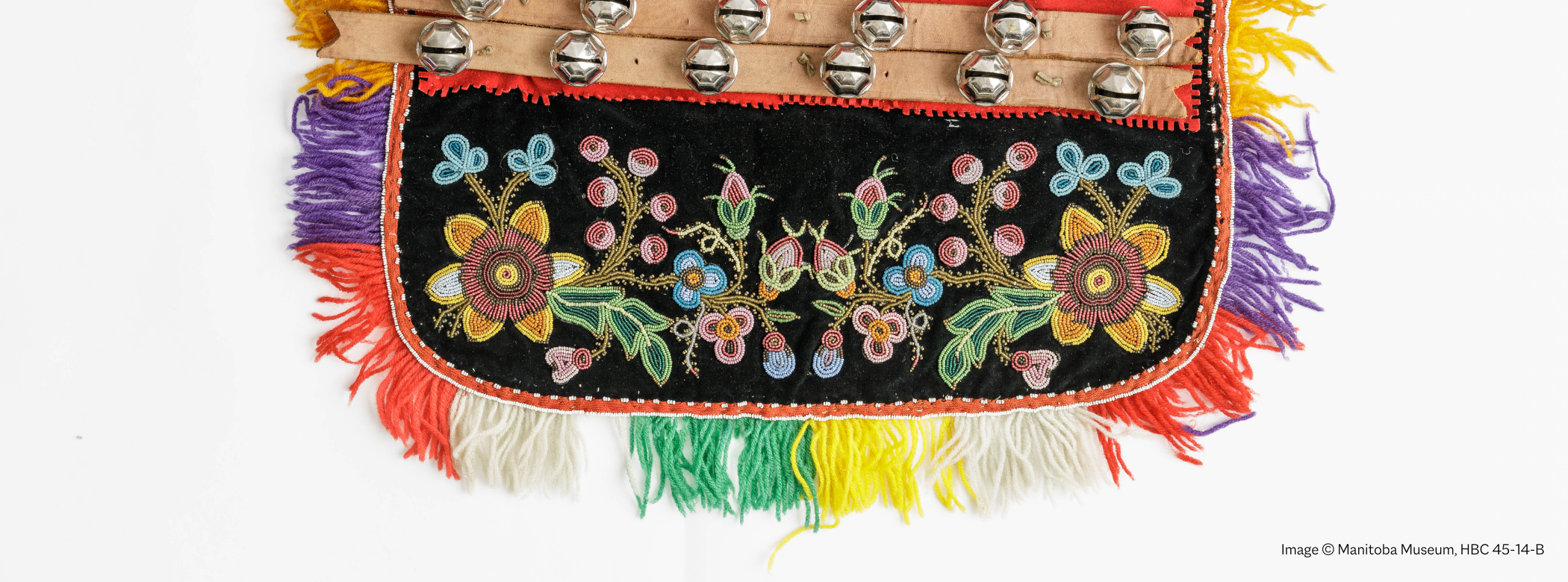 Close up on a half of a beaded dog blanket. Detailed floral beading on a black background, with a colourful fringe along the edges, and two rows of bells down the centre.