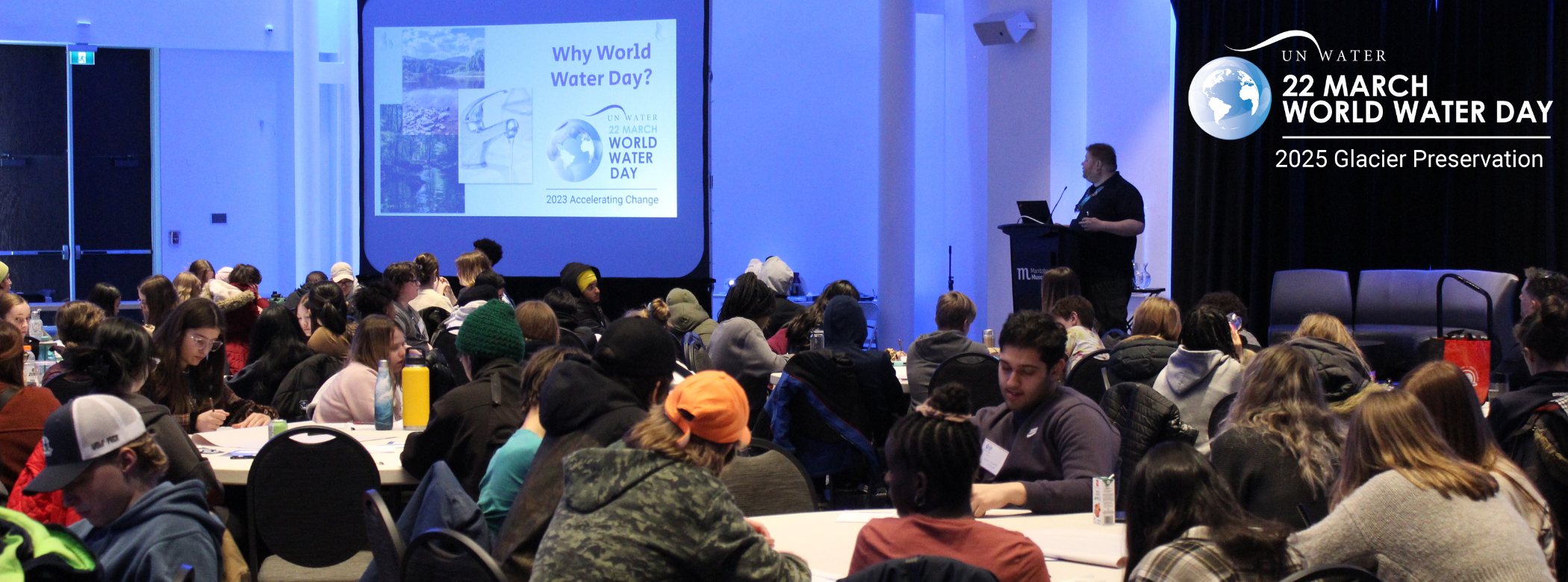 A large room with groups of students sitting around tables. In the back of the photo, a Museum staff person stands in front of a podium at the front of the room next to a presentation screen with 