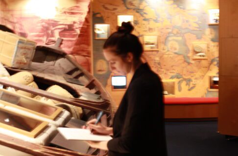 An individual standing in front of a large York Boat on display in the Hudson's Bay Museum Collection Gallery. They are holding a notepad and writing something on it.