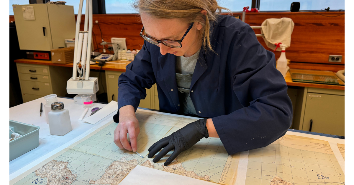 An individual wearing a navy blue lab coat leans over a paper map with a scalpel in one hand. Their other hand, in a black glove, carefully holds the paper in place.