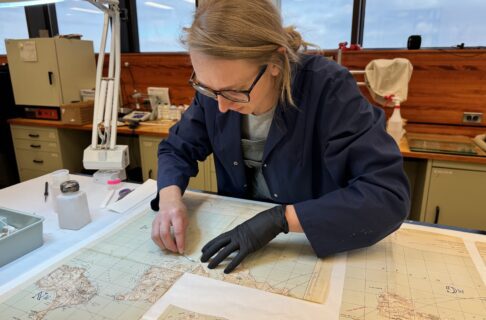 An individual wearing a navy blue lab coat leans over a paper map with a scalpel in one hand. Their other hand, in a black glove, carefully holds the paper in place.