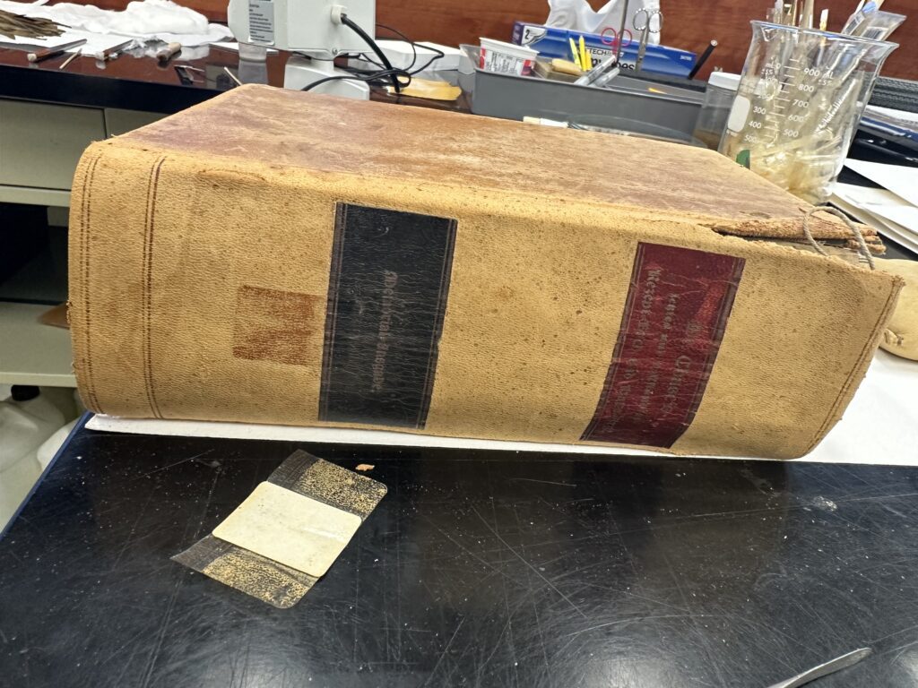 Large leather book with a red and black label on the spine. Book is sitting flat on a black table.