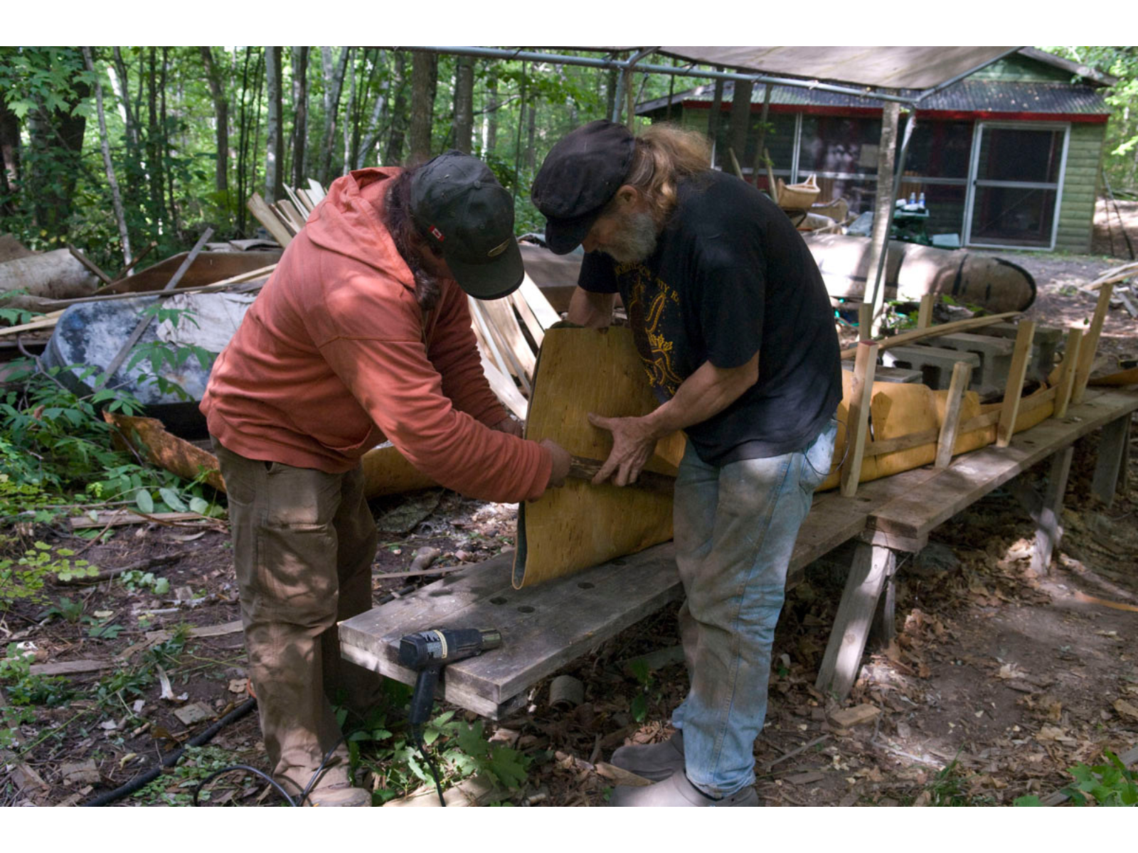 Two individuals working together to wrap and place birch bark strips around the frame of a canoe.