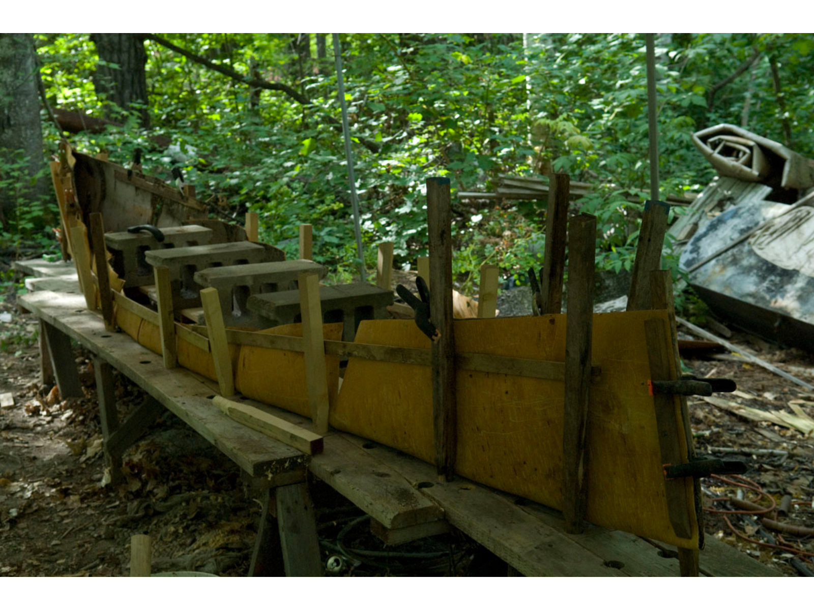 Wooden braces spiked into planks bracing the rough shape of a canoe, holding the birchbark in place, with cinderblocks in the middle weighing it down.