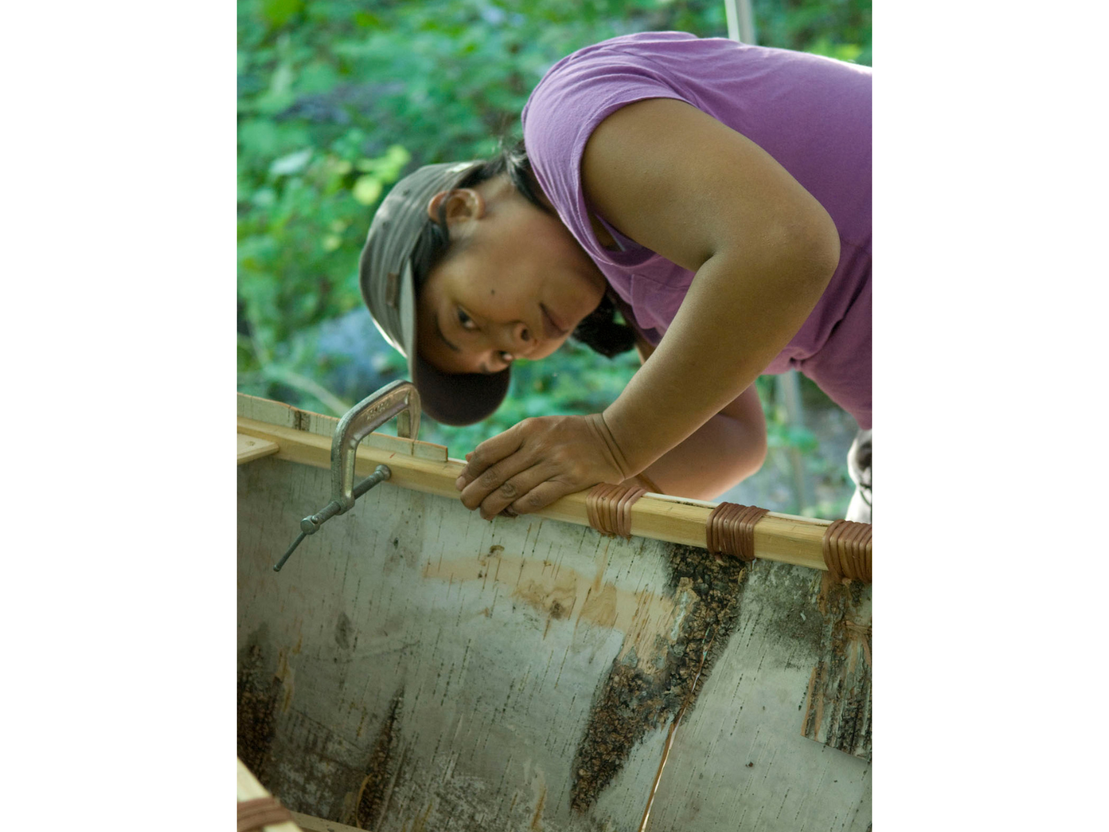 An individual leaning over at the side of a in-construction canoe, sewing along the upper part of the frame.