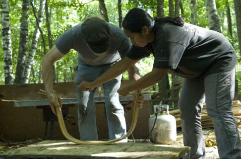 Two individuals working together to bend a strip of wood into a rough, wide horseshoe shape.