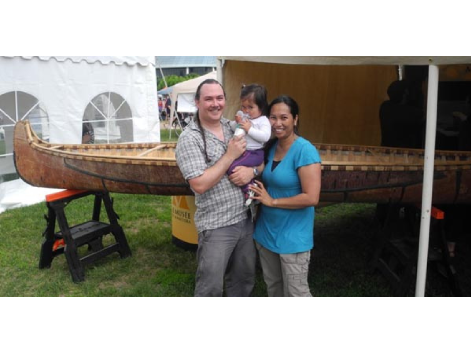 A couple standing together holding a toddler as they all stand in front of a canoe raised on saw horses until a canopy.