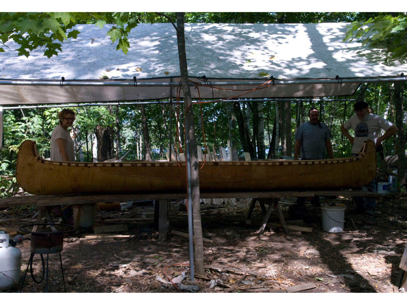 A birch bark canoe on sawhorses under an open sided tent. Three individuals stand at the ends of the canoe.