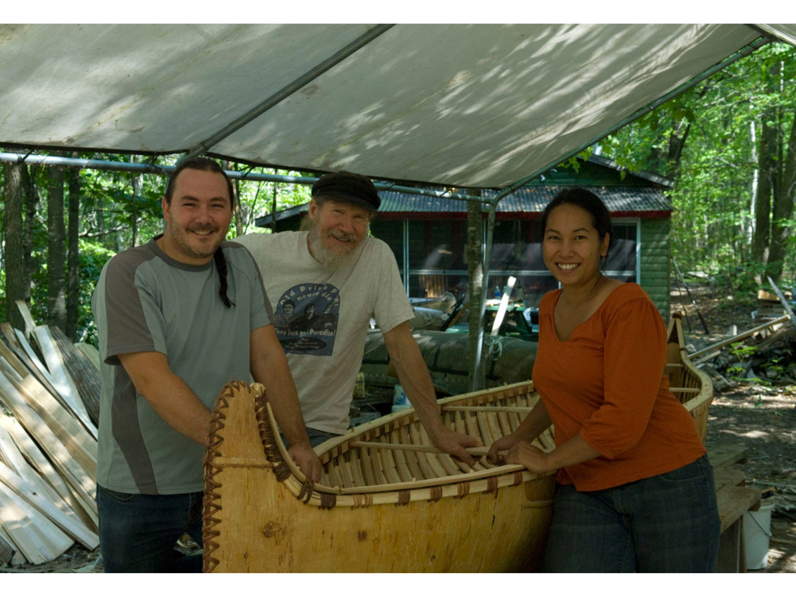 Three smiling individuals posing around a birch bark canoe supported on sawhorses under an open-sided tent.