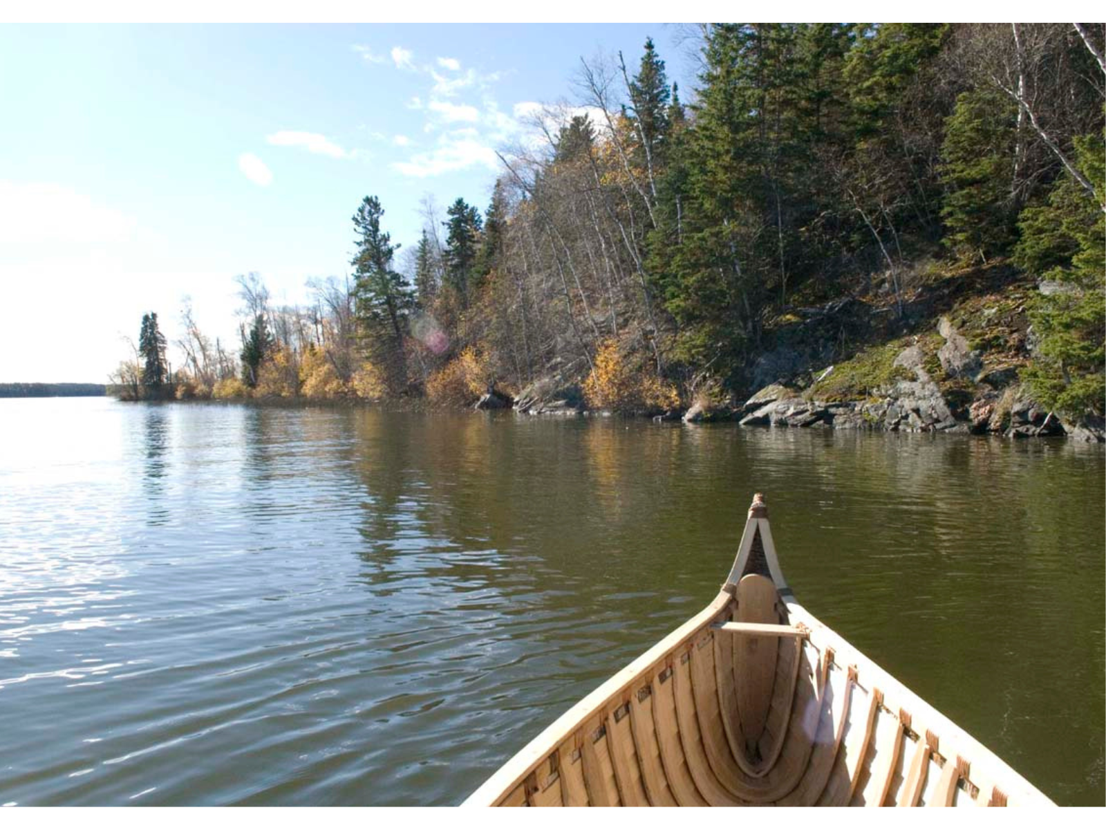 The view over the bow of a canoe on a body of water. To the right is a rocky and treed bank.