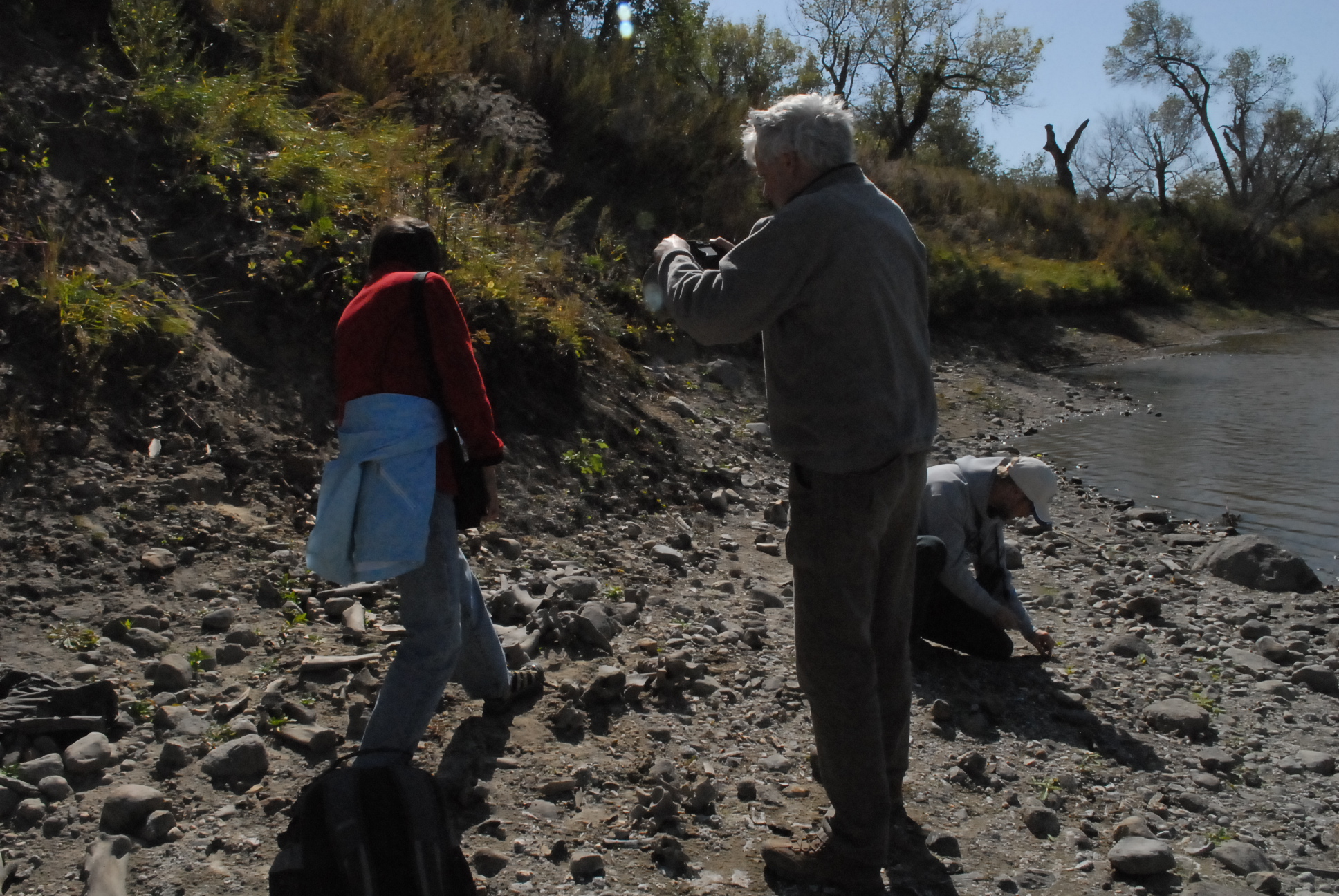 Three individuals on a river bank, one crouched looking at something on the ground, one mid-step, and one standing.