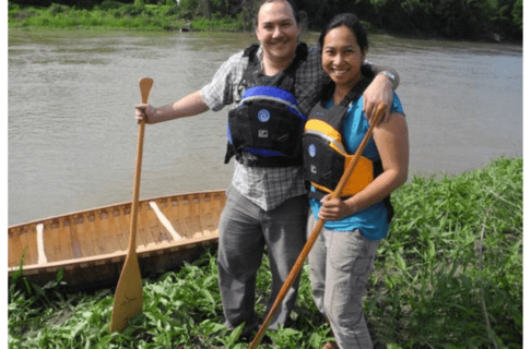 A couple with their arms around each other wearing life jackets and holding paddles standing on a bank next to a canoe in a river.