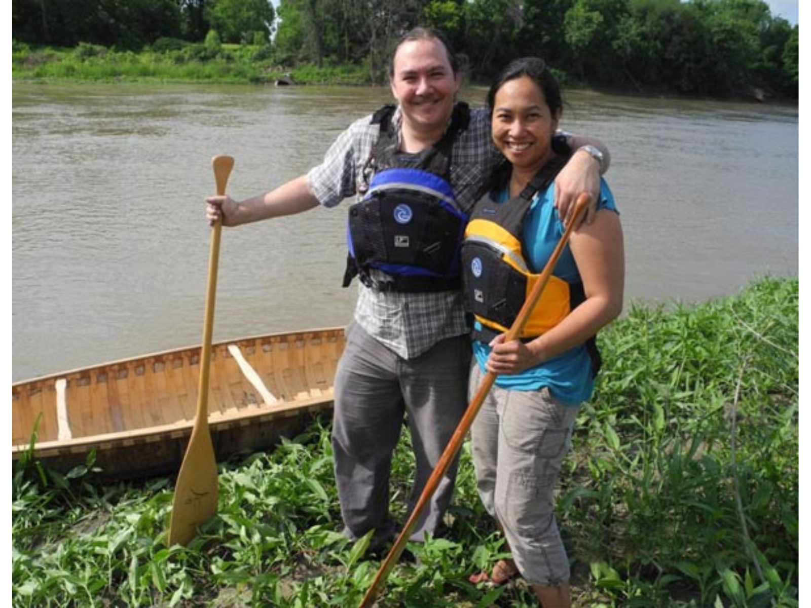 A couple with their arms around each other wearing life jackets standing on a bank next to a canoe in a river.