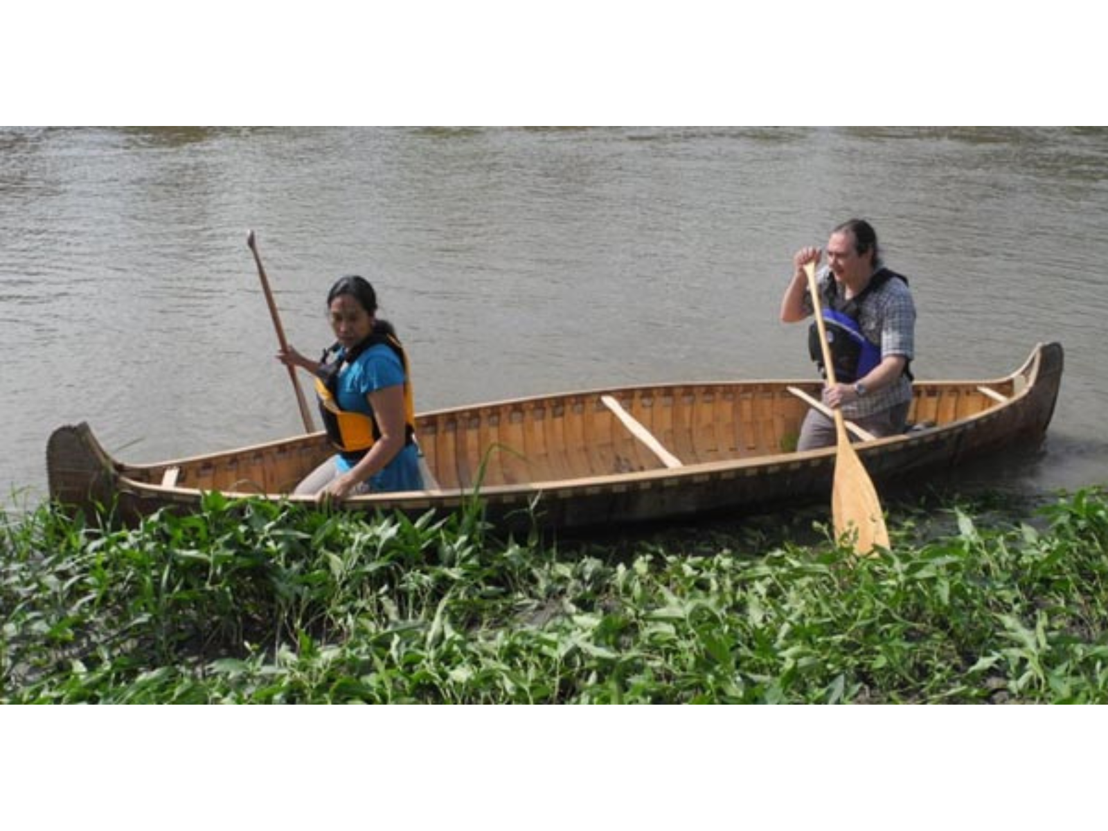 Two individual seated either end of a canoe, each using paddles to push off from a river bank.