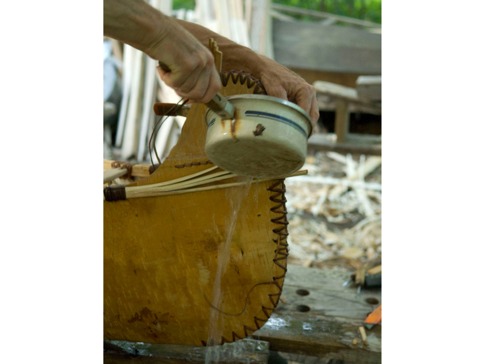 Hands reach into frame holding a small saucepot and pouring water onto the exterior end of a birchbark canoe.