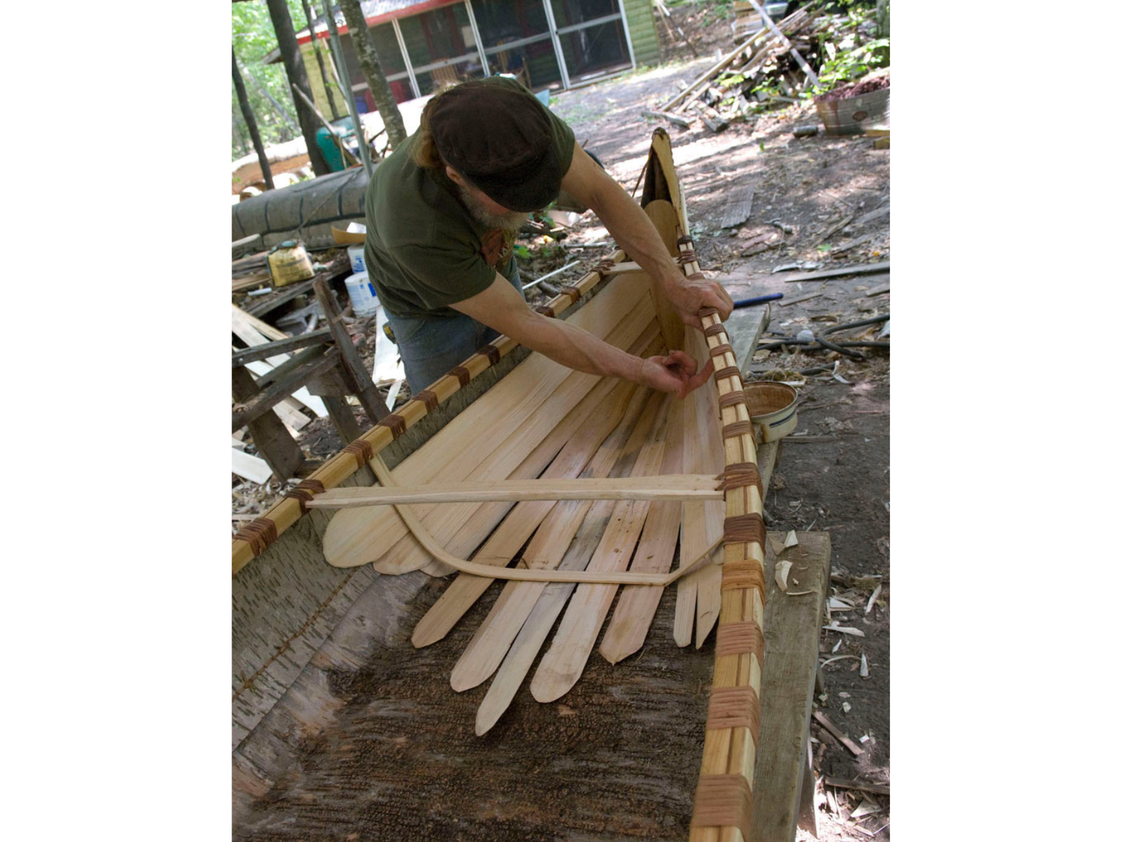 A partially constructed birchbark canoe, with an individual placing planking along the base of the canoe.