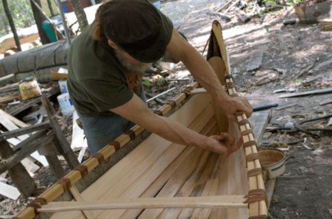 A partially constructed birchbark canoe, with an individual placing planking along the base of the canoe.