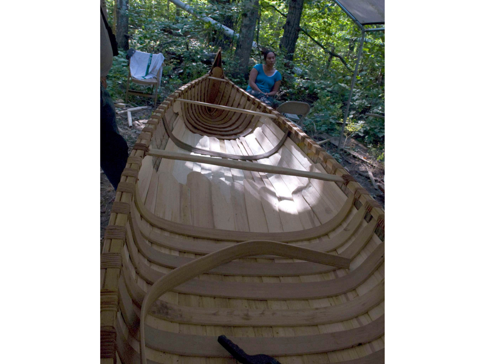 Looking down a birchbark canoe under an open-sided tent. Ribs are placed along half of the canoe so far. At the far end an individual rests in a lawn chair.