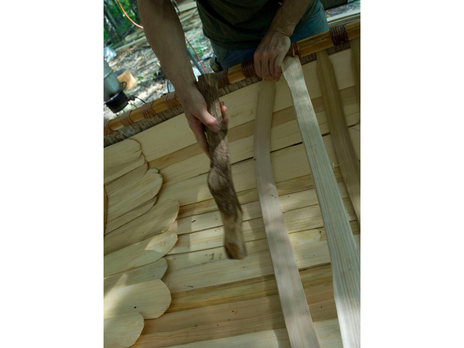 A hand reaches into frame holding a tick stick of iron wood as a mallet to place the ribs in the base of the canoe.