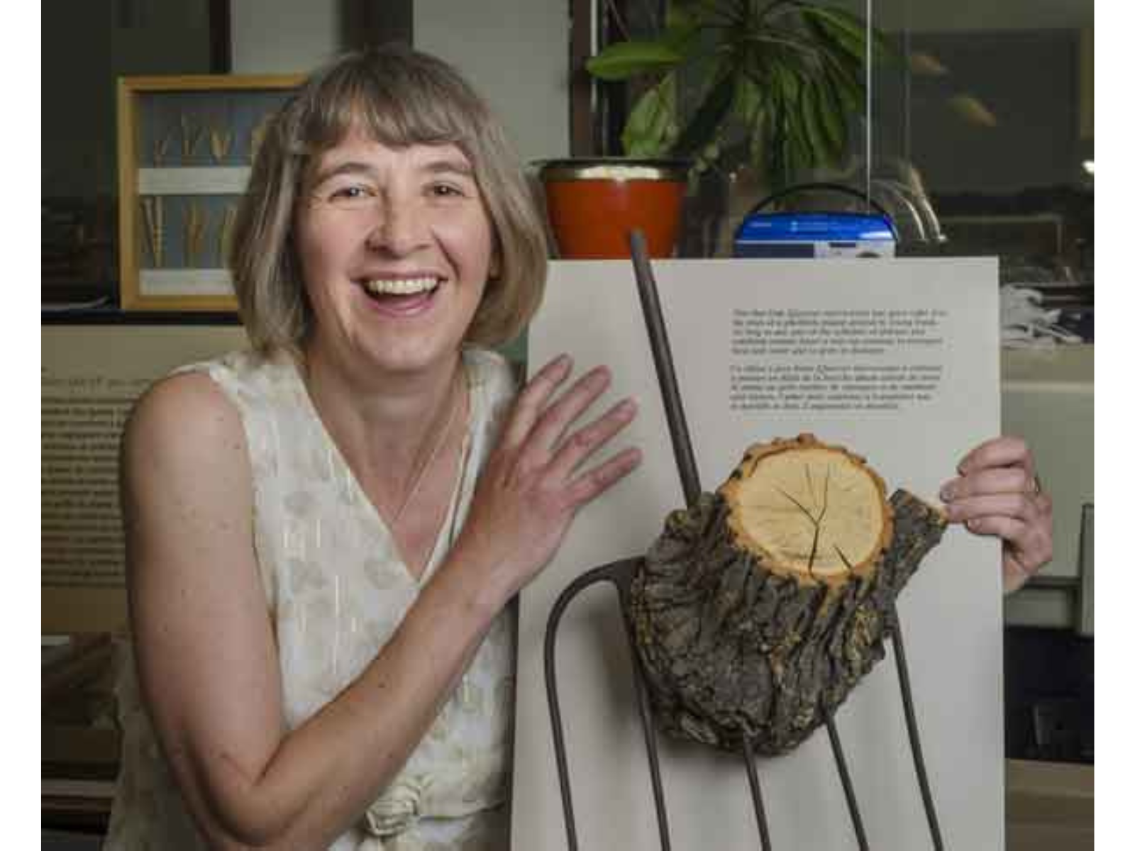 A smiling individual holds up a piece of tree trunk mounted on a display board, growing around the metal end of a pitchfork.