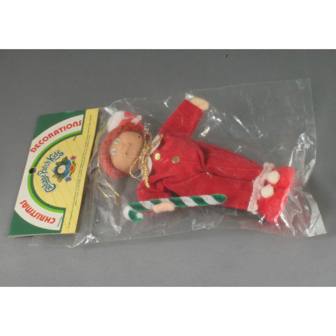 A small Cabbage Patch Doll wearing a red onsie pyjama set, red shoes with white bobbles, and a red bonnet. The ornament is still in the original packaging.